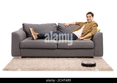 Young man sitting on a sofa and a robotic vacuum cleaner vacuuming the carpet isolated on white background Stock Photo
