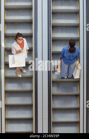Graphic top down view at two women riding escalator in opposite directions while shopping in mall, focus on woman wearing mask looking up at camera, copy space Stock Photo