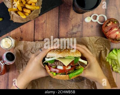 Person holding a burger with fries, onion rings and tomato on wooden table Stock Photo