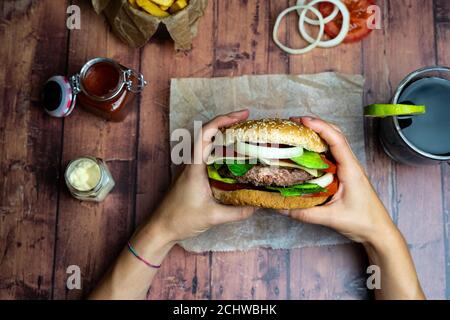 Person holding a burger with fries, onion rings and tomato on wooden table Stock Photo