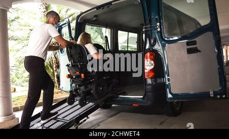 Woman on wheelchair going into vehicle using an accessible ramp with assistant helping. Stock Photo