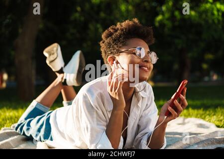 Portrait of happy smiling african woman lying outdoors in park on a grass and using mobile phone while listening music Stock Photo