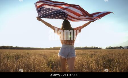 Young happy American girl running and jumping on 4th of July carefree with open arms over wheat field. Holding USA flag.