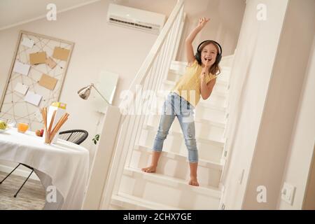 Little singer. Cute and happy caucasian girl wearing headphones standing on stairs and listening music, she is singing and having fun at home