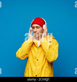Portrait of positive cool man holding headphone listening music with closed eyes, male isolated over blue color background. Bright casual wear, yellow cloak and red hat Stock Photo