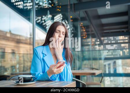 Tired woman bored and yawning while is working on the phone, watching media in a mobile sitting at table at breakfast on a balcony at home or on a res Stock Photo