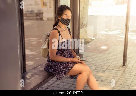 Young Hispanic female wearing a medical mask and sitting down to wait for her bus Stock Photo