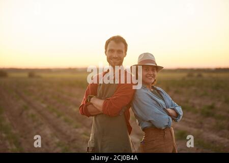 Waist up portrait of young farmers couple posing back to back while standing in field at sunset and smiling at camera, copy space Stock Photo