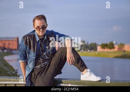 Man is looking out through sunglasses while sitting on the railing of the bridge. Male 27 years old during touristic trip over Europe. Stock Photo