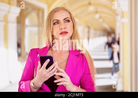 Woman in pink blazer is using smartphone in the shopping center. Middle aged female with phone in hands is going across the  arched gallery of the dep Stock Photo