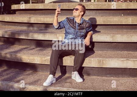 Smiling man 27 years old is sitting on the beton stair and taking selfie during walk over the city. Young male is making pictures of himself using sma Stock Photo