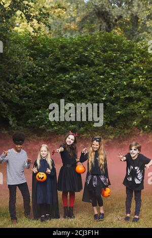 Vertical full length portrait of spooky children wearing Halloween costumes outdoors with colored red smoke in background, copy space Stock Photo