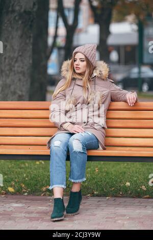 Young woman in autumn clothes sits on a bench in a city park. The woman is dressed in a stylish jacket with fur Stock Photo
