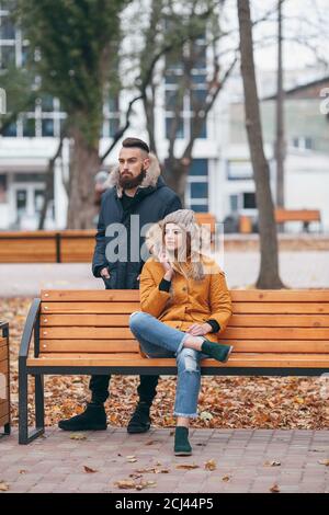A guy and a girl are resting on a bench in an autumn park. A loving couple in jackets sits on a bench in the main park Stock Photo