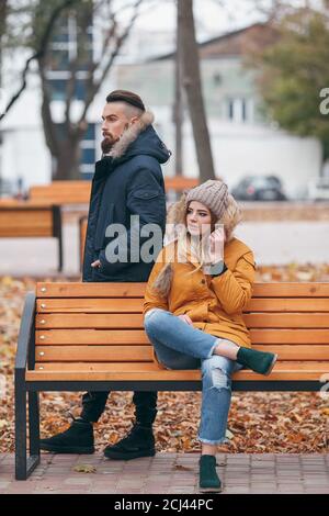 A guy and a girl are resting on a bench in an autumn park. A loving couple in jackets sits on a bench in the main park Stock Photo