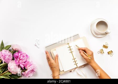 Well-groomed woman hands is writing in diary with stationery supplies set onside and peonies bouquet Stock Photo