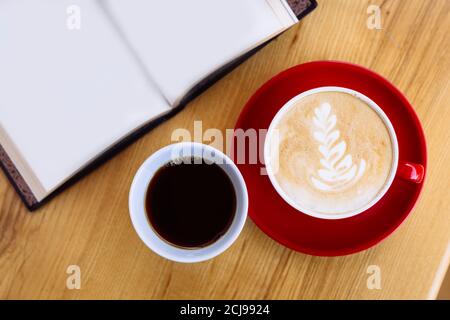 A mug of fresh aromatic coffee and a cappuccino coffee mug, an open book next to it, on a wooden table. Flat lay