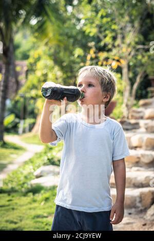 Front view of happy thirsty blond kid boy drinking dark tea from plastic bottle standing outside in summer park nature background. Healthy and Stock Photo