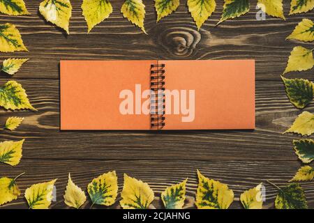 Notepad with orange pages in a frame of yellow leaves on a wooden background. Creative flat lay autumn composition. Top view, copy space. Mock up. Stock Photo