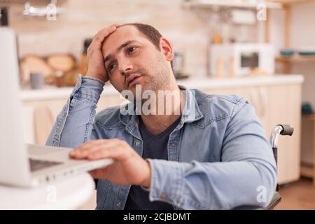 Stressed businessman with walking disability in wheelchair using laptop in kitchen. Disabled paralyzed handicapped man with walking disability integrating after an accident. Stock Photo