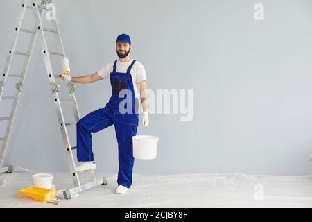 Smiling painter standing with paint roller and bucket in apartment for repairing and looking at camera Stock Photo