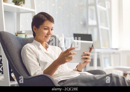 Happy lady watching videos on tablet sitting in cozy armchair in the living room Stock Photo