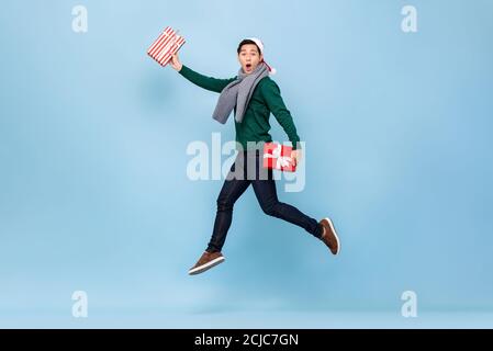 Asian man in Christmas sweater jumping and holding gift boxes isolated in light background Stock Photo