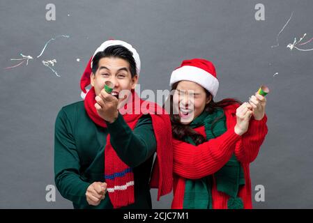 Cute excited Asian couple pulling party poppers celebrating Christmas in isolated studio gray background Stock Photo