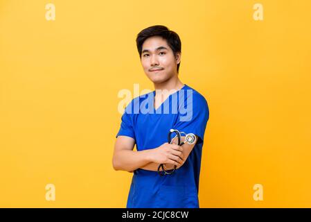 Young handsome Asian male doctor smiling and doing arms crossed gesture isolated on yellow background Stock Photo