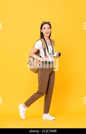Full length portrait of smiling cute young Asian woman tourist with camera waling on isolated yellow studio background Stock Photo