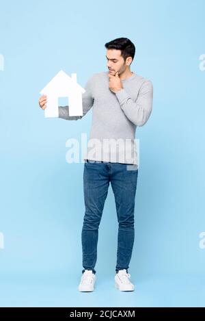 Full length portrait of thoughtful Caucasian man holding house model cutout and thinking in light blue isolated studio background