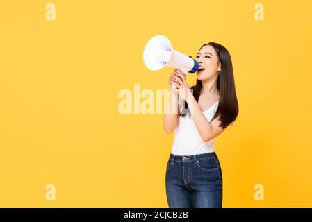 Portrait of smiling cheerful young pretty Asian woman holding megaphone making announcement in isolated studio yellow background Stock Photo