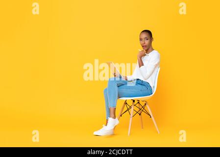 Thinking young African American woman sitting and using tablet computer with stylus pen on isolated yellow background Stock Photo