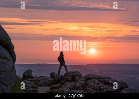 Man standing on Haytor rocks at sunrise silhoutted against a bright orange sky Stock Photo