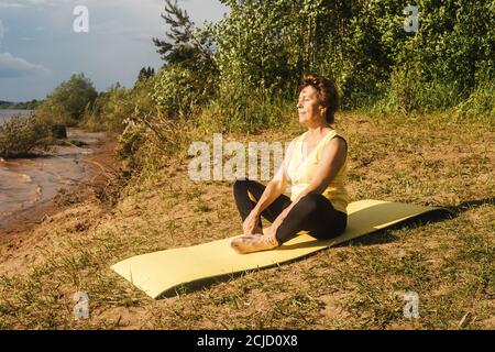 Senior woman making yoga near river in summer. Front view. Stock Photo