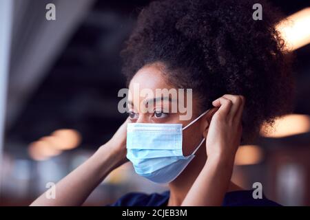 Female Doctor In Scrubs Putting On Face Mask Under Pressure In Busy Hospital During Health Pandemic Stock Photo