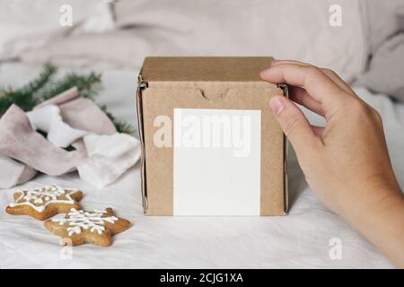 Christmas gift box label mockup scene. Closeup of womans hand holding gift box. Velvet ribbons, gingerbread cookies and spruce tree branches on linen Stock Photo