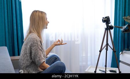 Young woman in casual clothes records herself on camera on tripod. Side view. Cheerful female blogger recording video at home Stock Photo