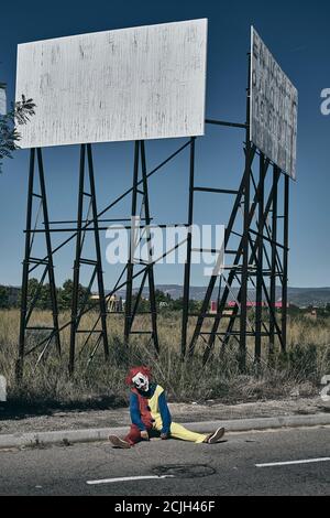 a scary evil clown, wearing a yellow, red and blue costume outdoors, sitting on the ground on a rural road in front of the rusty structure of an aband Stock Photo