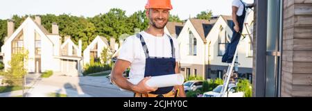 Horizontal concept of builder holding blueprint while working with colleague on roof of building Stock Photo
