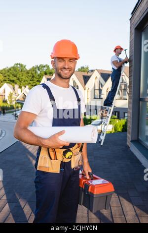 Selective focus of builder in tool belt holding blueprint and toolbox near colleague on roof Stock Photo