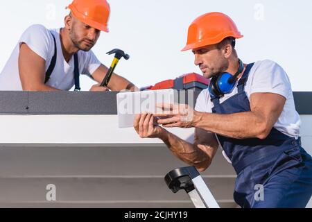 Repairmen in overalls and helmets using digital tablet on rooftop of house Stock Photo
