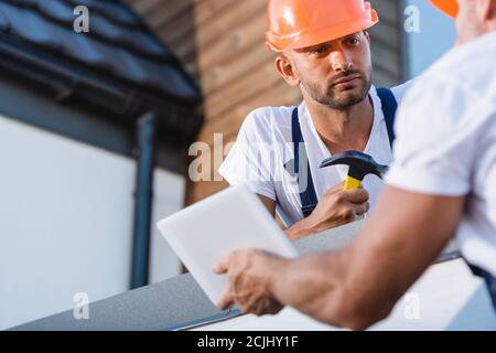 Selective focus of builder with hammer looking at colleague with digital tablet on roof of building Stock Photo