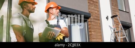 Panoramic shot of builders in workwear using digital tablet and looking away near building Stock Photo