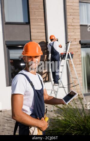 Selective focus of builder holding digital tablet while colleague working on ladder near building Stock Photo