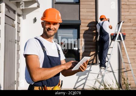 Selective focus of builder with digital tablet looking at camera while colleague working on ladder near house at background Stock Photo