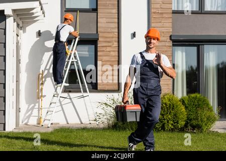 Selective focus of handyman with toolbox showing ok at camera while standing on lawn near building Stock Photo