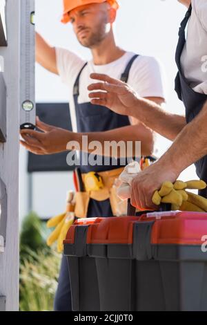 Selective focus of workman holding gloves and toolbox while colleague using spirit level near house Stock Photo