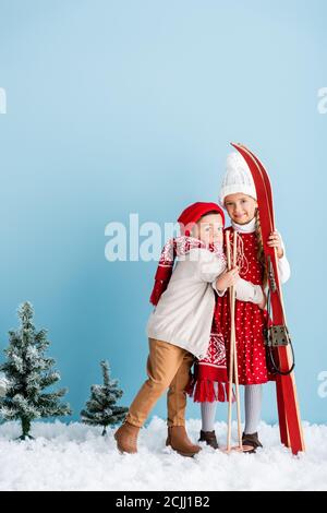 kid in hat and scarf hugging sister with ski poles and skis while standing on blue Stock Photo