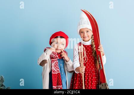 kid in winter outfit with ski poles and skis standing near brother in hat on blue Stock Photo